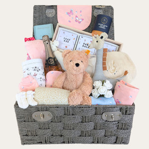 Baby shower gifts for girl packed in a large hamper basket. Presents include pink baby clothes set and skincare for mum.