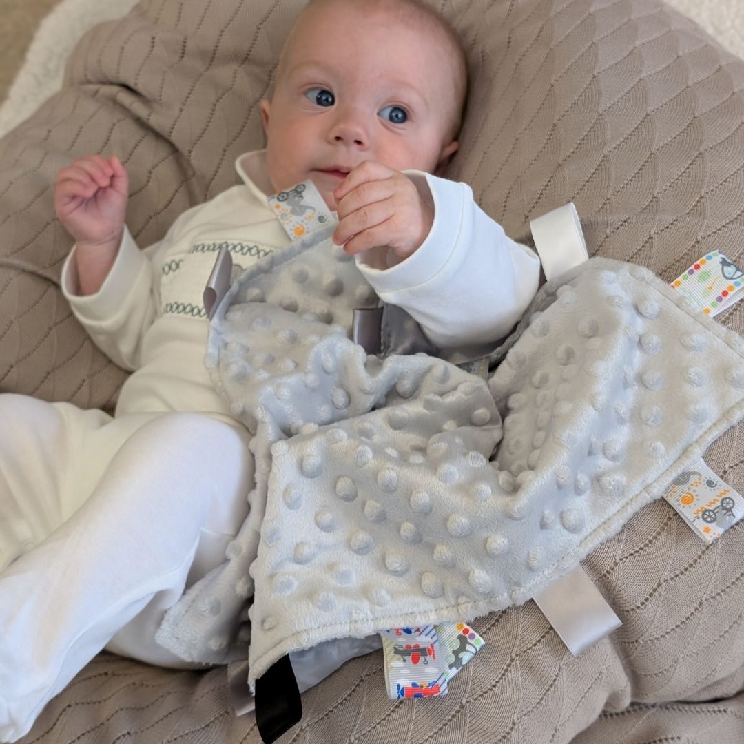 Baby holding a Soft Grey Taggie Comforter Blanket With Ribbons.