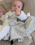 Baby holding a Soft Grey Taggie Comforter Blanket With Ribbons.