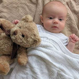 Baby covered in a white cellular baby blanket with a teddy bear.